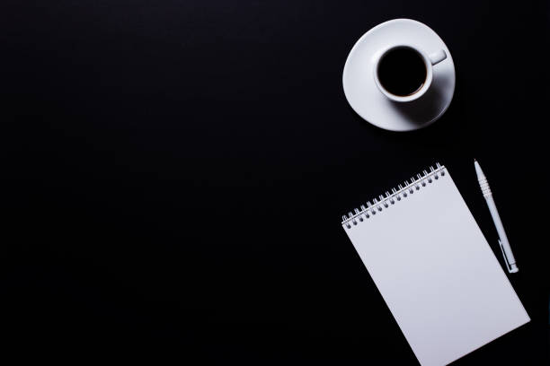 istock White cup of coffee, blank notebook and white pen on a black background. Business concept closeup top view 1335675821