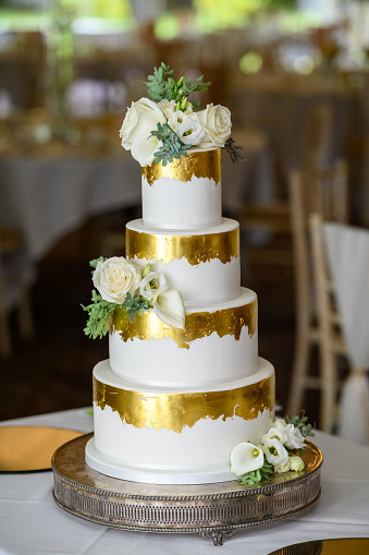 A bride and groom, who are wheelchair users that have just got married, at their evening reception in a wedding venue in Morpeth, North East England. They are out of focus, looking at the camera while smiling behind their wedding cake. The cake is in focus and has figurines of the happy couple made out of icing on top of it.