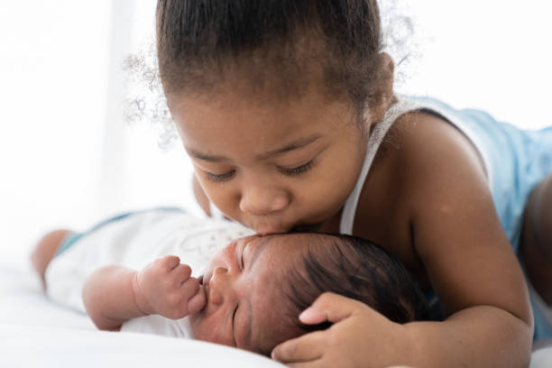 cute african american little girl kissing on newborn baby cheek on white bed at home. little girl takes care of infant baby with kindly - mother baby new kissing imagens e fotografias de stock