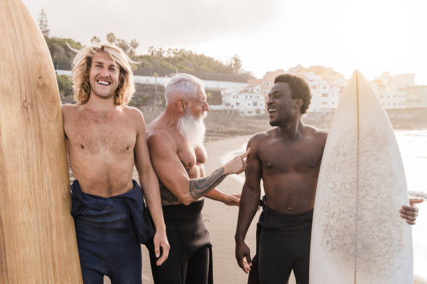 divers amis surfeurs tenant des planches de surf après une séance de sports nautiques extrêmes avec la plage en arrière-plan - focus sur l’homme âgé - opposition party photos et images de collection