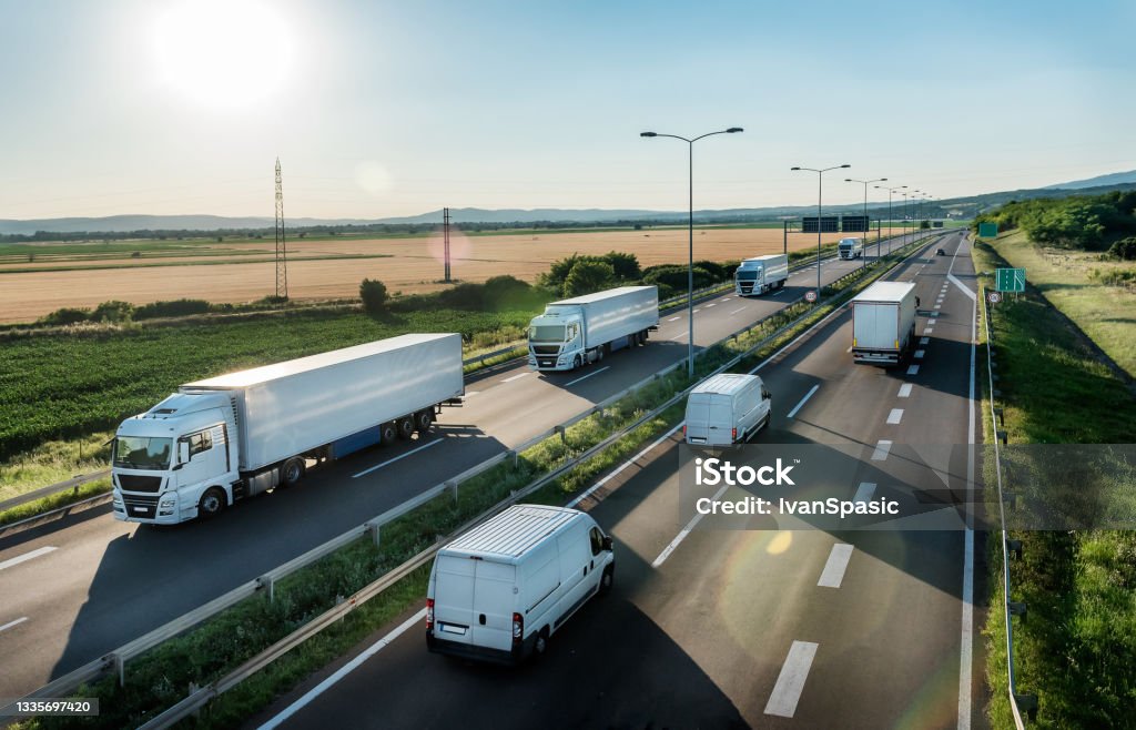Highway Transit photo with Convoy of transportation trucks Convoy or caravans of transportation trucks passing vans and truck on a highway on a bright blue day. Highway transit transportation with lorry trucks and vans Convoy Stock Photo