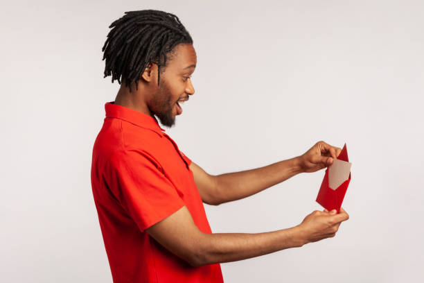 vista lateral do homem positivo com dreadlocks vestindo camiseta estilo casual vermelho, carta de abertura em envelope vermelho, segurando cartão de boas-vindas e se sente feliz. - opening mail envelope greeting card - fotografias e filmes do acervo