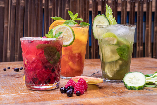 Front view of 3 fresh fruit juices in glasses on a wooden table, there are cucumber and peach slices on the table, with raspberries and blueberries, glasses garnished with mint leaves, lime and orange slices.