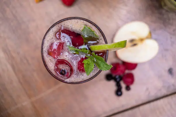 Top view of a glass of red coloured fresh fruit and veggie juice with some ice cubes and berries in the glass, on a wooden table, some berries and half an apple are seen blurry on the table.