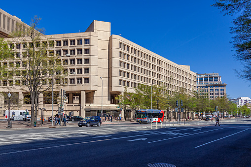 Federal Bureau of Investigation (FBI) Headquarters on Pennsylvania Avenue, Washington DC, USA. Clear Blue Sky, Street, Passersby, Cars and Green Trees are in the image. Wide angle lens.