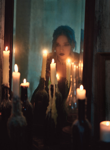 Indoors portrait of beautiful goth girl among candles. Blue-haired gothic woman looking into the old dirty mirror. Young witch. Vintage style