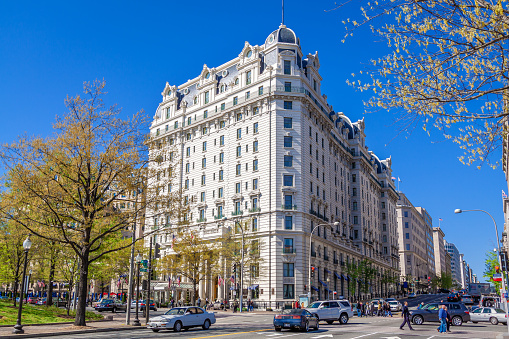 The Willard InterContinental Washington  (Willard Hotel), is located at Pennsylvania Avenue NW, Washington DC, USA. It was built in architectural style known as a luxury Beaux-Arts. Clear Blue Sky, Street, Passersby, Cars and Green Trees are in the image. Wide angle lens.