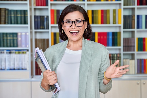 Portrait of positive smiling middle aged woman teacher, psychologist, mentor, therapist, counselor, business woman looking at camera. Female in glasses with textbooks tablet, library office with books