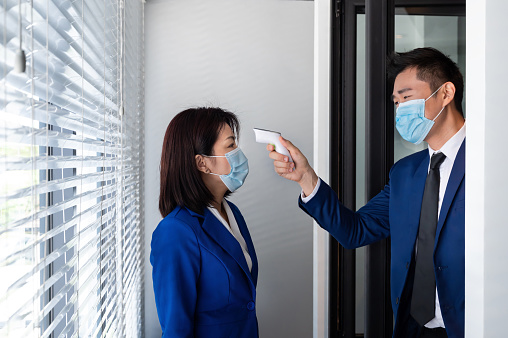 Office employee checking body temperature of a colleague with digital thermometer before allow her entering the office. The office new normal with wear a facemask, social distancing, body temperature check and disinfection practice during the global Coronavirus COVID-19 pandemic.