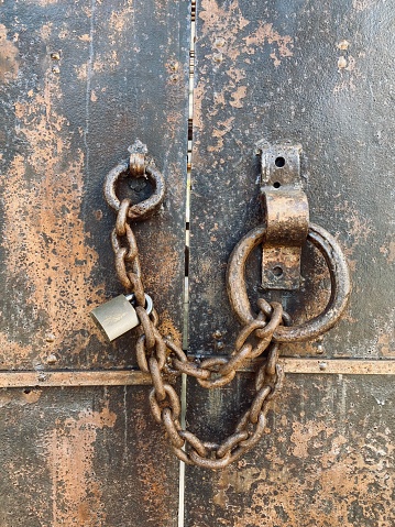 Closeup photo of an ancient, stained, weathered rusty metal ring door handle and security chain with a modern padlock.