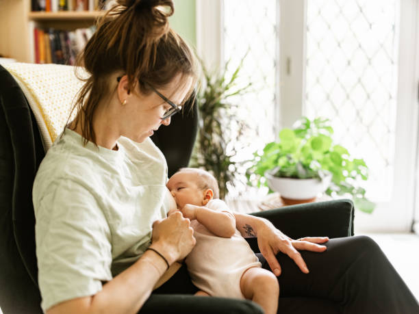 Young mother with baby at home Young mother with baby at home. Young mother dressed in casual clothes wearing glasses, breastfeeding 3 month old baby girl in her arms inside nursery bedroom of private home. BREAST MILK stock pictures, royalty-free photos & images