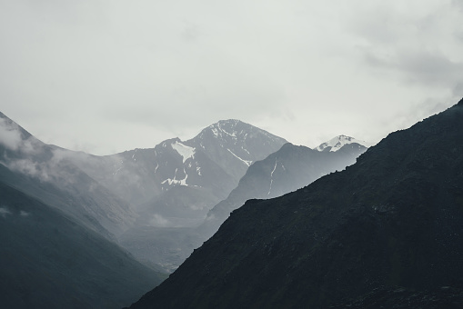 Dark atmospheric landscape with black mountains silhouettes among rainy low clouds. Gloomy mountain scenery with rainy clouds on silhouette of mountain top in overcast weather. Peak in gray cloudy sky