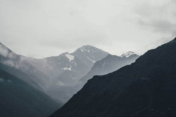Photo of Dark atmospheric landscape with black mountains silhouettes among rainy low clouds. Gloomy mountain scenery with rainy clouds on silhouette of mountain top in overcast weather. Peak in gray cloudy sky