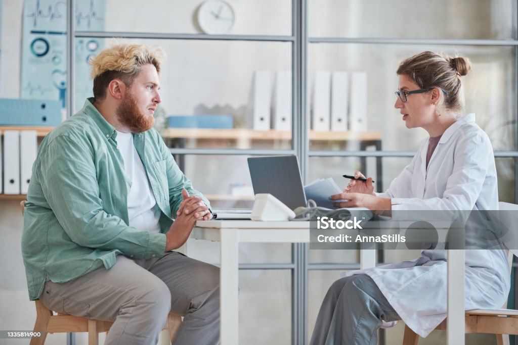 Man talking to the doctor at office Overweight man sitting at the table together with doctor and talking about his health during his visit Overweight Stock Photo