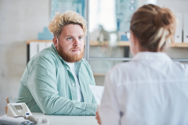 Man visiting his nutritionist Young overweight man sitting at the table at hospital he visiting a doctor for having a nutrition recommendations overweight stock pictures, royalty-free photos & images