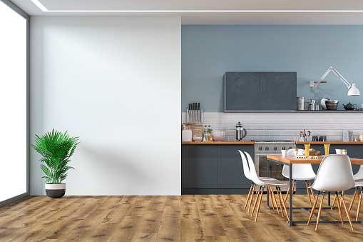 Empty anthracite modern kitchen on hardwood floor with appliances, full dining table, chairs in front of white metro tiled wall partly light blue plaster wall background. A potted plant in front of a large white plaster wall background with copy space and windows on a side. 3D rendered image.
