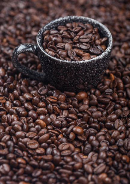 Black ceramic cup with fresh raw coffee beans inside beans background. Macro