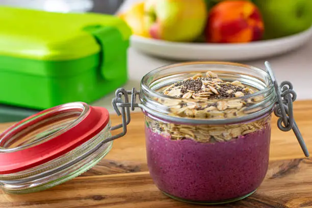 Take out food  for breakfast, lunch or snack for work or for school with a fresh blueberry yogurt topped with oatmeal and chia seeds served in a glass jar with led on kitchen table background