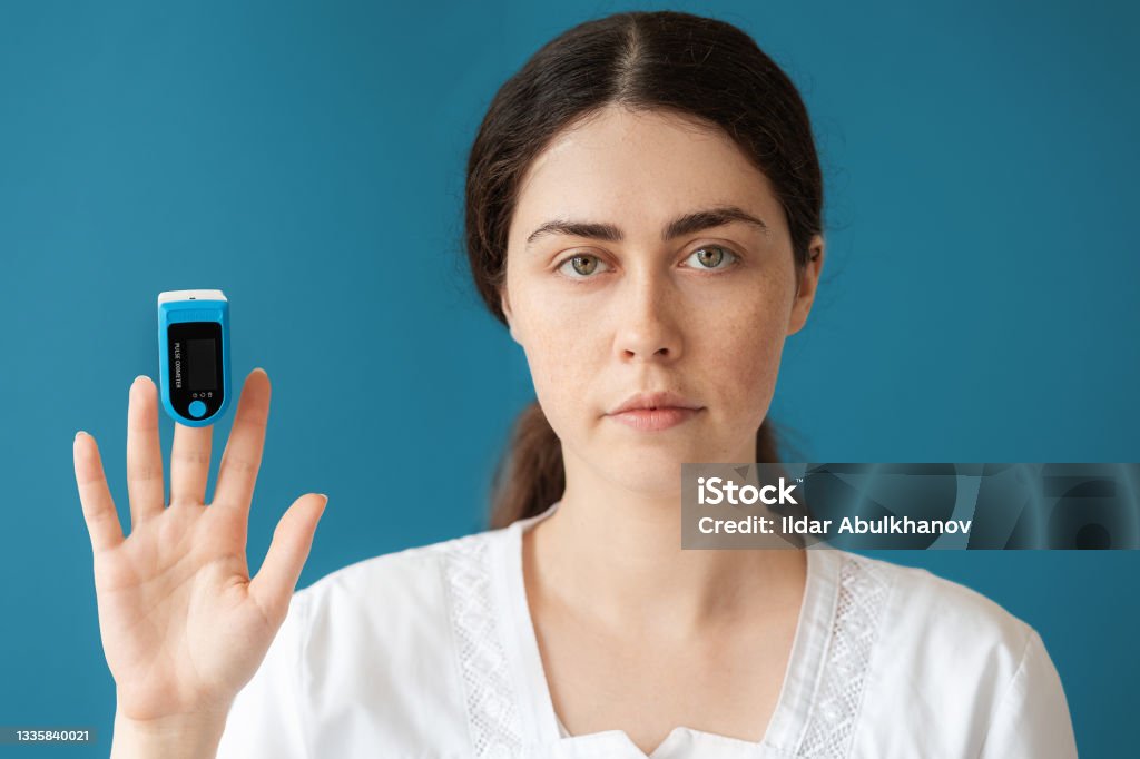 Portrait of woman doctor shows a pulse oximeter on her index finger. Blue background. The concept of check health and medical insurance Portrait of woman doctor shows a pulse oximeter on her index finger. Blue background. The concept of check health and medical insurance. Pulse Oximeter Stock Photo