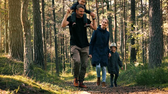 active young family walks on the forest trail with children. son sitting on dad shoulders