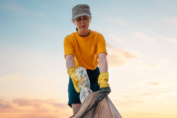 Portrait of female volunteer in rubber gloves puts a dirty plastic bottle in bag with garbage. Sunset sky on the background. Bottom view. The concept of envirinmental pollution and ecology Portrait of female volunteer in rubber gloves puts a dirty plastic bottle in bag with garbage. Sunset sky on the background. Bottom view. The concept of envirinmental pollution and ecology. littering stock pictures, royalty-free photos & images