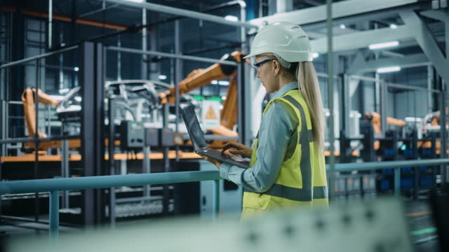 Car Factory: Female Automotive Engineer Wearing Hard Hat, Standing, Using Laptop. Monitoring, Control, Equipment Production. Automated Robot Arm Assembly Line Manufacturing Electric Vehicles. Medium