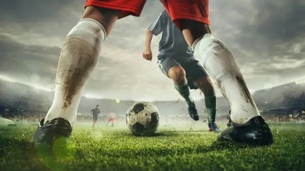 Close up legs of football or soccer players during football match at a stadium filled with spectators, fans. Moment of attacking. Concept of sport, competition, winning, action, motion, overcoming.