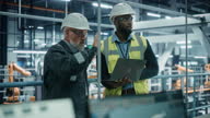 istock Car Factory: Project Manager and Automotive Engineer in Hard Hat, Talking, Using Laptop. Monitoring, Control, Equipment Production. Automated Robot Arm Assembly Line Manufacturing Electric Vehicles 1335860559