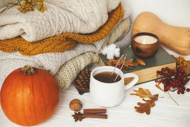 Photo of Hello autumn, cozy slow living. Pumpkin, cup of tea, cozy sweaters, autumn leaves, burning candle and vintage book on white wooden background in room. Happy Thanksgiving
