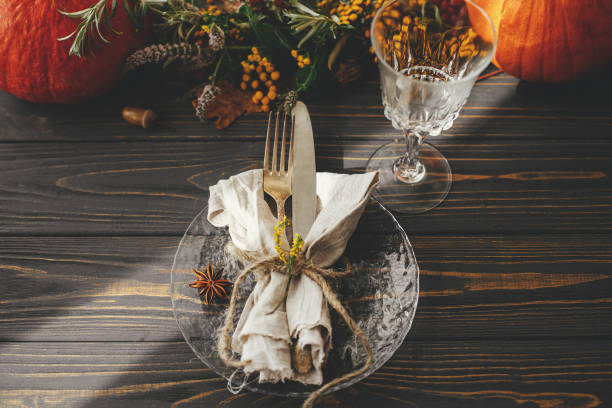 istock Thanksgiving dinner table setting. Modern plate with vintage cutlery, linen napkin, herb and glass on wooden table with pumpkins and autumn flowers arrangement in sunlight. Rustic catering 1335868111