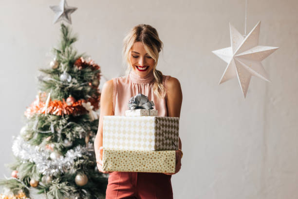 Happy Woman Holding a Christmas Presents in her Hands Cheerful smiling female standing next to the decorated Christmas tree and holding wrapped gift boxes in hands christmas present stock pictures, royalty-free photos & images
