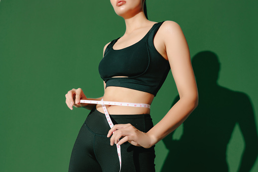 Crop close up body of sporty woman measuring her thin waist with a tape measure isolated on green background. Dynamic movement. Strength and motivation.