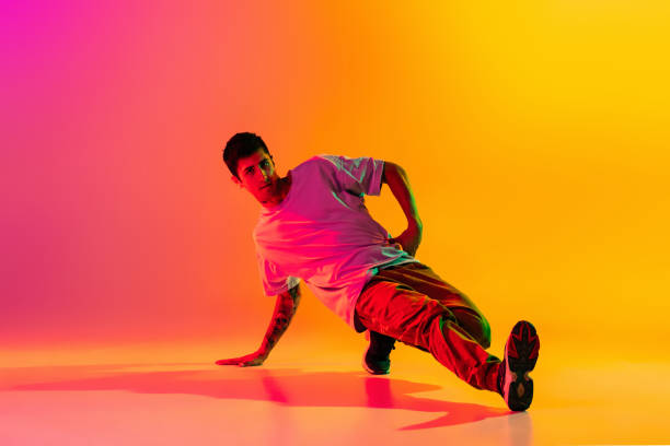 istock Solo performance. Young man, break dancing dancer ptacticing in casual clothes isolated over gradient pink yellow background at dance hall in neon light. 1335872765