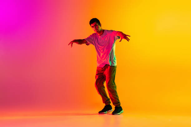 istock Solo performance. Young man, break dancing dancer ptacticing in casual clothes isolated over gradient pink yellow background at dance hall in neon light. 1335872829