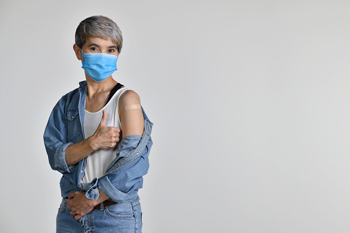 Happy middle aged Asian woman vaccinated showing thumbs up at adhesive plaster bandage on her arm after coronavirus antiviral injection isolated on white background