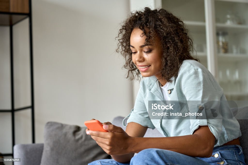 Smiling African American woman using mobile phone sitting on sofa at home. Smiling happy young African American woman holding cell phone sitting on sofa at home, using online mobile apps technology doing online shopping, dating in application, checking social media, texting. Subscription Stock Photo