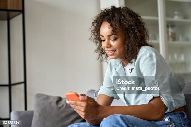 Smiling African American Woman Using Mobile Phone Sitting On Sofa At Home Stock Photo - Download Image Now