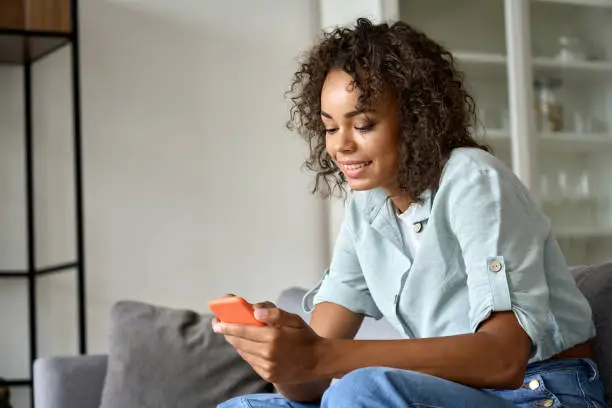 Photo of Smiling African American woman using mobile phone sitting on sofa at home.