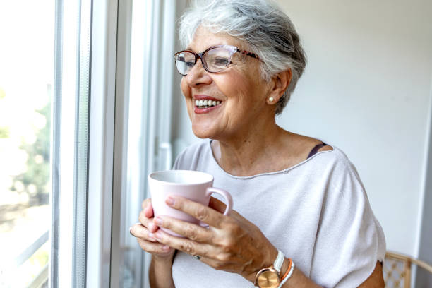 istock Charming elderly woman posing by window and smiling while drinking coffee from a pink cup. 1335881341