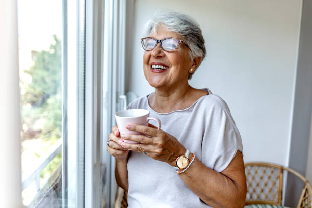 istock Charming elderly woman posing by window and smiling while drinking coffee from a pink cup. 1335881540