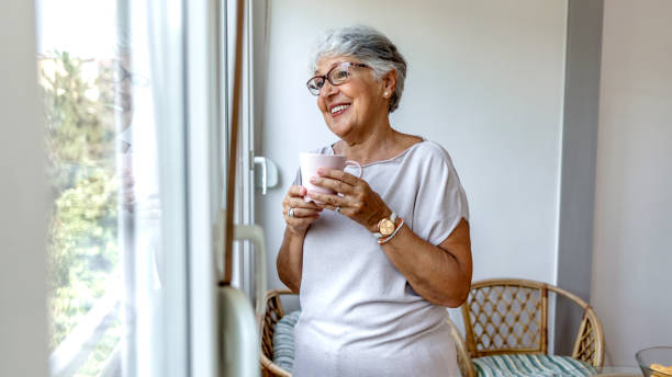 una mujer pensativa y feliz mirando por la ventana mientras sostiene una taza de café, copia espacio. - pensive senior adult looking through window indoors fotografías e imágenes de stock