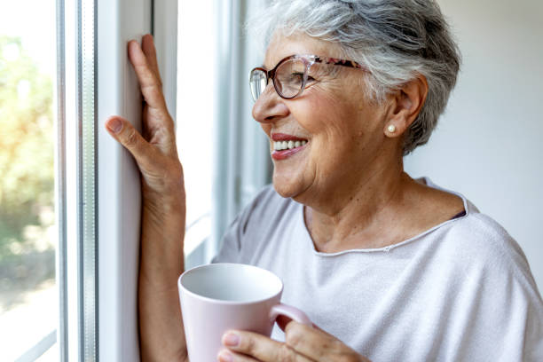 istock Charming elderly woman posing by window and smiling while drinking coffee from a pink cup. 1335882112