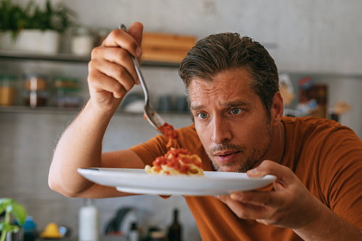 Focused handsome young man preparing and tasting spaghetti with tomato sauce