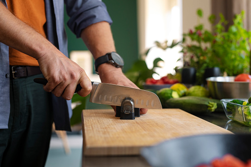 Unrecognizable handsome young man sharpening kitchen knife on wooden board