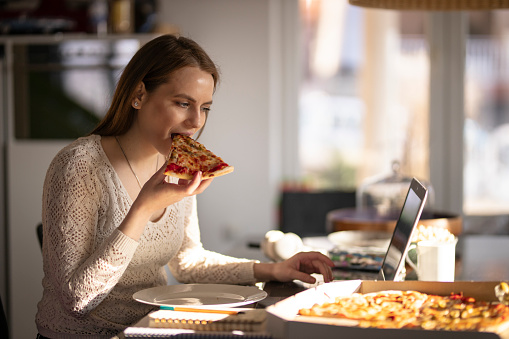 Young woman eating take-out pizza, using a laptop, sitting in the living room, and working from home