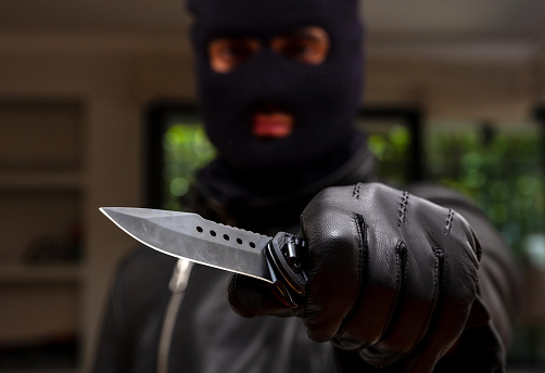Burglar with balaclava, holding a knife in left hand, armed robbery concept. Robber threaten with a dagger, blur indoor background, closeup view.