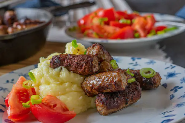 mediterranean dish with spicy meat rolls, serbian cevapcici served with mashed potatoes and a tomato salad with olive oil and chives on a plate