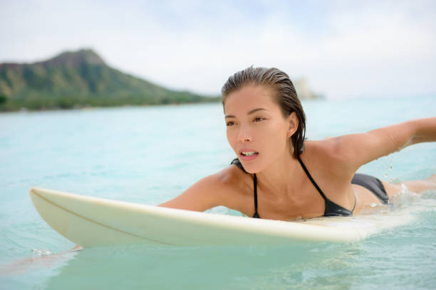 Surfing surfer girl paddle for surf on surfboard. Female bikini woman living healthy active water sports lifestyle on Hawaiian beach. Asian Caucasian model on Waikiki Beach, Oahu, Hawaii.