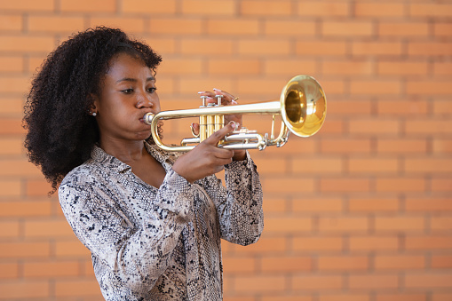Afro american woman with curly hair playing the trumpet on a brick wall background. Selective focus.