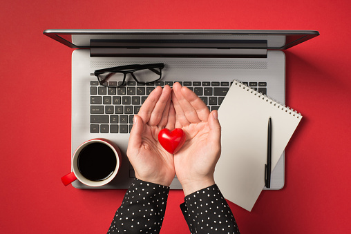 Overhead photo of grey laptop notepad pen glasses cup of coffee and hands holding a red heart isolated on the red backdrop